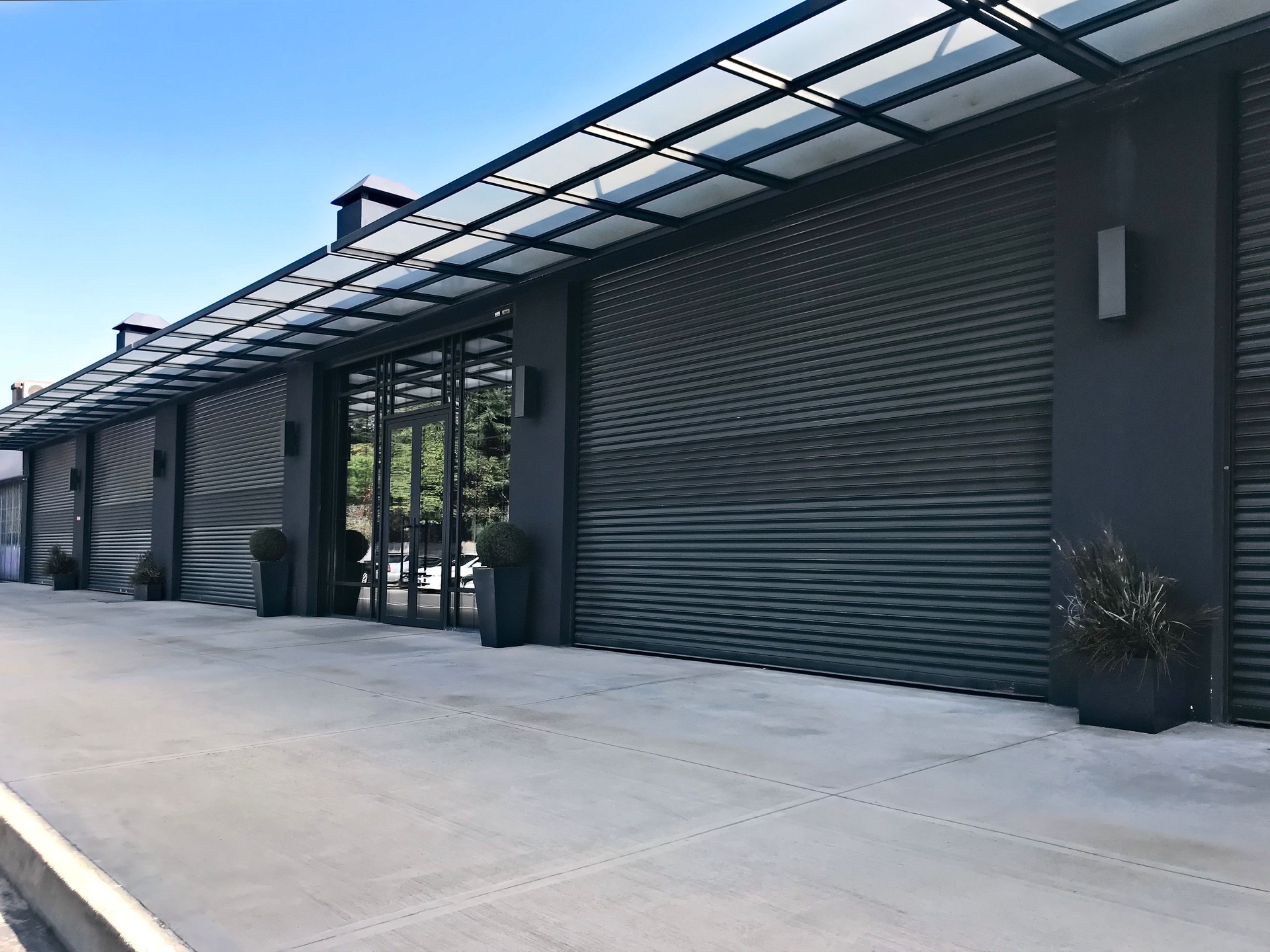 Modern commercial metal building with roll-up doors, glass storefront, and sidewalk, representing a pre-engineered steel structure by Texas Metal Buildings. Modern commercial metal building with roll-up doors, glass storefront, and sidewalk, representing a pre-engineered steel structure by Texas Metal Buildings.